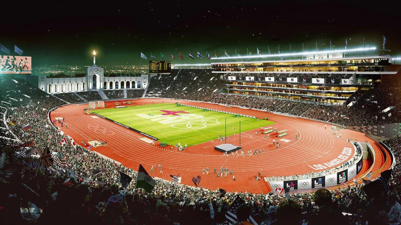 Rendering of the LA Memorial Coliseum during the 2028 Olympics at night. The stadium is filled with spectators, and flags from various countries are waving in the crowd. The field and the track surrounding it are branded with the Olympic rings and the LA28 logo. The iconic Olympic torch is lit atop the coliseum's peristyle. The stadium screens display live scoring and competition footage, and the stadium lights illuminate the entire scene.