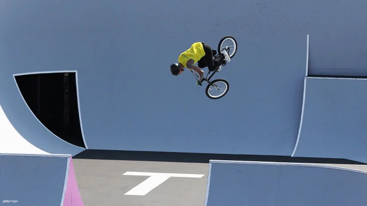 A BMX rider is performing an aerial trick high above the ramps in a skatepark. Wearing a bright yellow shirt, black pants, and a black helmet, the rider is captured mid-air, sitting on the BMX bike with their body bent forward. The skatepark's ramps and surfaces are light blue, gray and white.