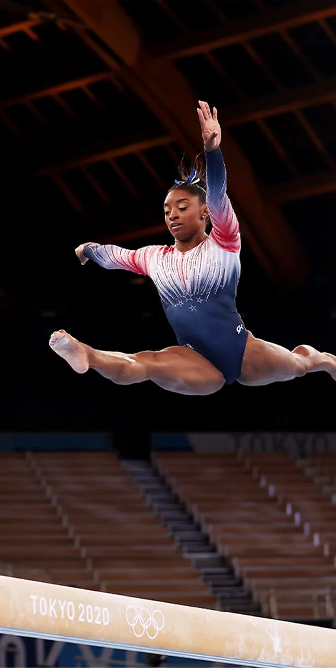 Photograph of Simone Biles doing a leap on a beam with her arms extended while wearing a red white and blue leotard