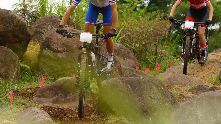 Two cyclists navigate a rocky terrain on mountain bikes during a competitive race. The cyclist in the foreground, wearing a blue and white uniform with stripes on the sleeves, is maneuvering over large boulders, while the cyclist behind them, dressed in a red and black outfit, follows closely. Pink markers are placed on the ground to indicate the race course, and lush greenery surrounds the rocky path. The focus is on the trail and their bikes.