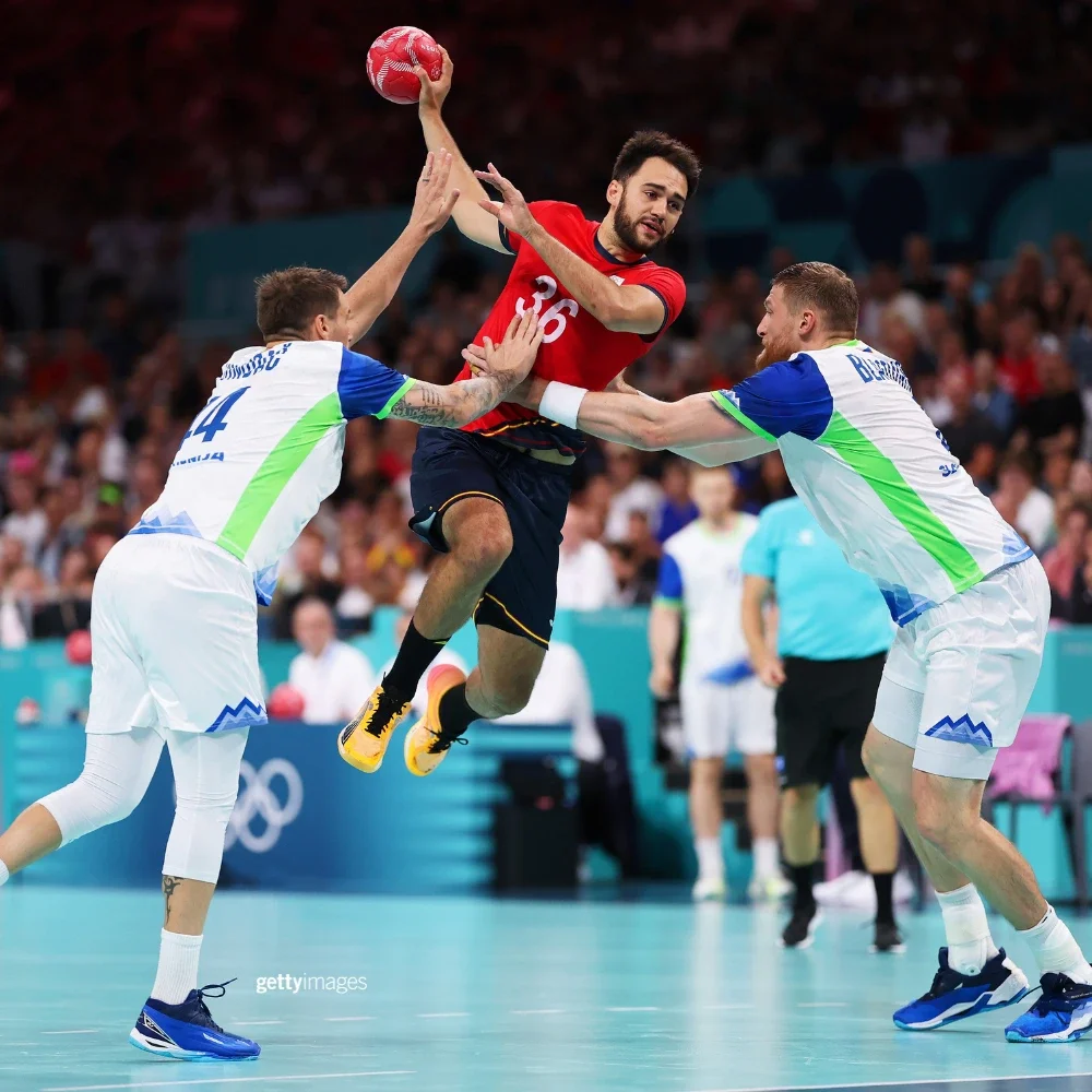 A photo of a handball player from Spain jumping in the air while holding a ball in his hand while two other players on the opposing team in blue and white uniforms try to defend the goal