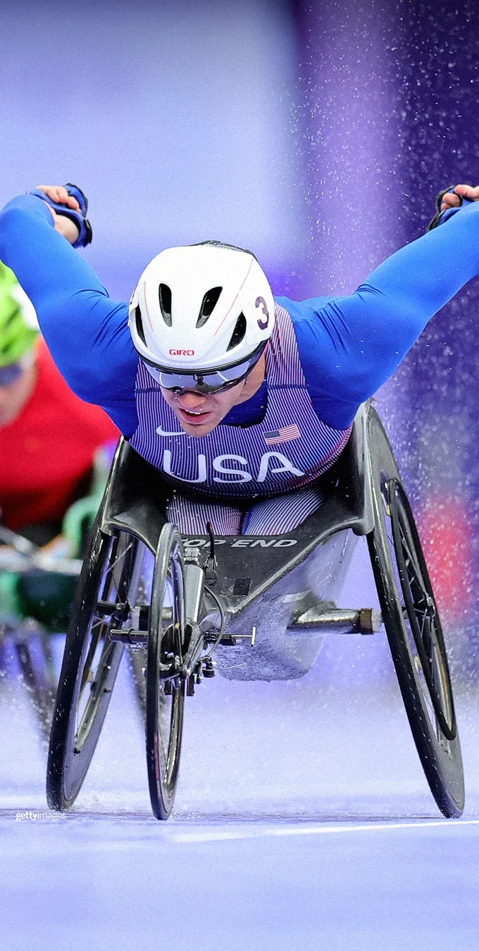 Cyclists pedaling in the Paralympic Games on handcycles, the cyclist in the forefront is wearing a blue Team USA long-sleeved top, a helmet and sunglasses