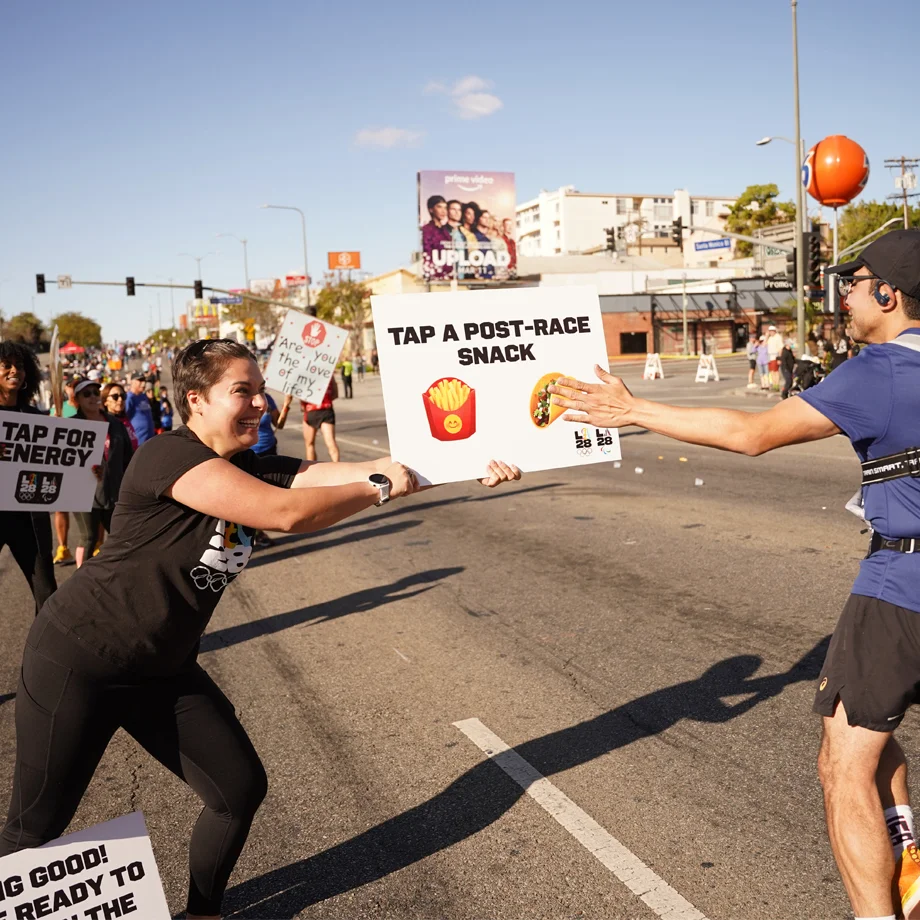 LA28 Employee holding a sign that says Tap A Post Race Snack which has a picture of fries and a taco and runner is touching the picture of the taco