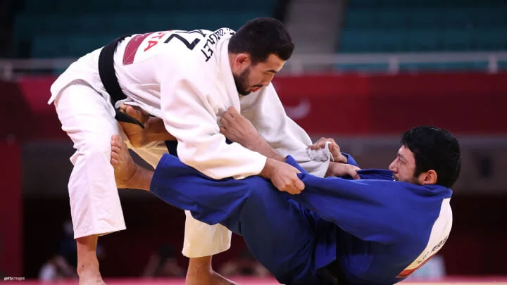 Dos judokas están involucrados en un combate de judo. El judoka a la izquierda viste un gi blanco y tiene agarre en el cuello y la manga del judoka a la derecha, quien usa un gi azul. El judoka con el gi azul está en el suelo, empleando su pierna derecha para presionar contra la cadera y pierna del judoka con el gi blanco, mientras sostiene el gi blanco con las manos.