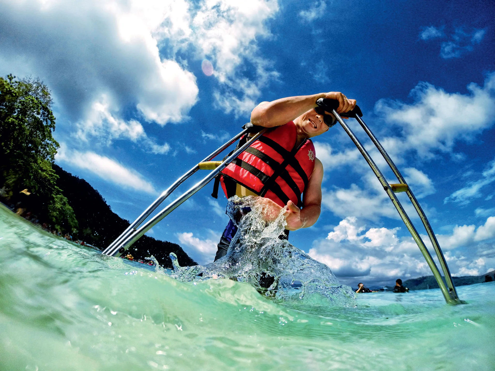 Photo of a man wearing sunglasses walking in the ocean with crutches and a life jacket