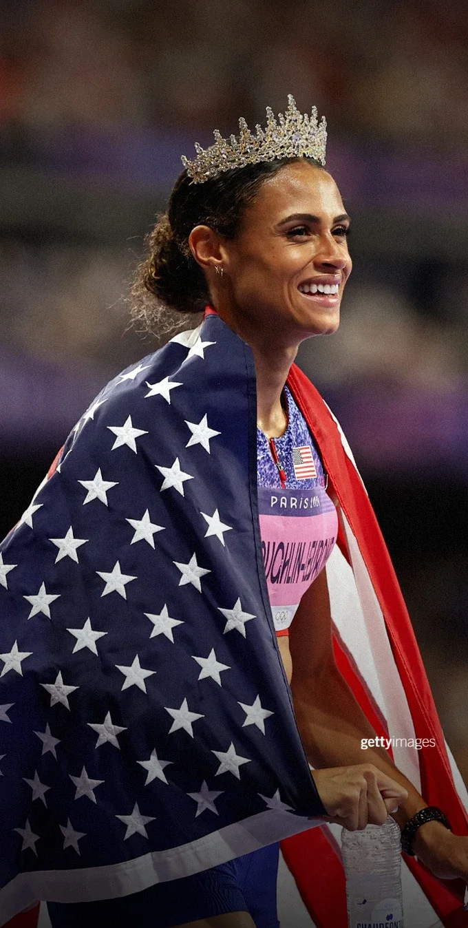 Photograph of Sydney McLaughlin-Levrone wrapped in an American flag with a crown on her head after winning a gold medal at the Paris 2024 Games