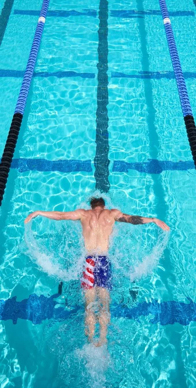 Photo of a male with a tattoo sleeve on his right arm, swimming in a pool with aquamarine colored water while wearing shorts with an American flag print.