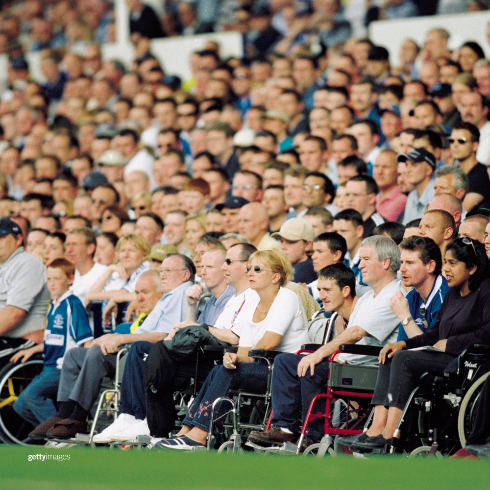 Spectators at a sport event with spectators in wheelchairs in the front row with grass on the filed in the foreground of the image. 