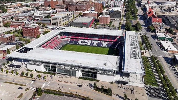 Aerial view of St. Louis Stadium with the empty stadium seats showing the word 'City' in the stands. Buildings from downtown St. Louis surround the stadium and a street is shown in the forefront of the image, in front of the stadium entrance.