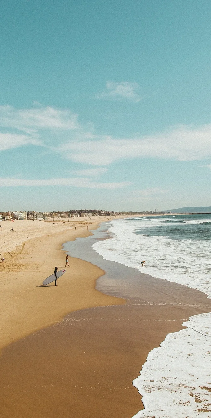 Photograph of a beach with tan sand and light blue waves with foam washing up on the shore. There is a sufer holding a surfboard standing on the sand and a woman watching a child play in the surf. In the background homes line the sandy shore and people are sitting in the sand