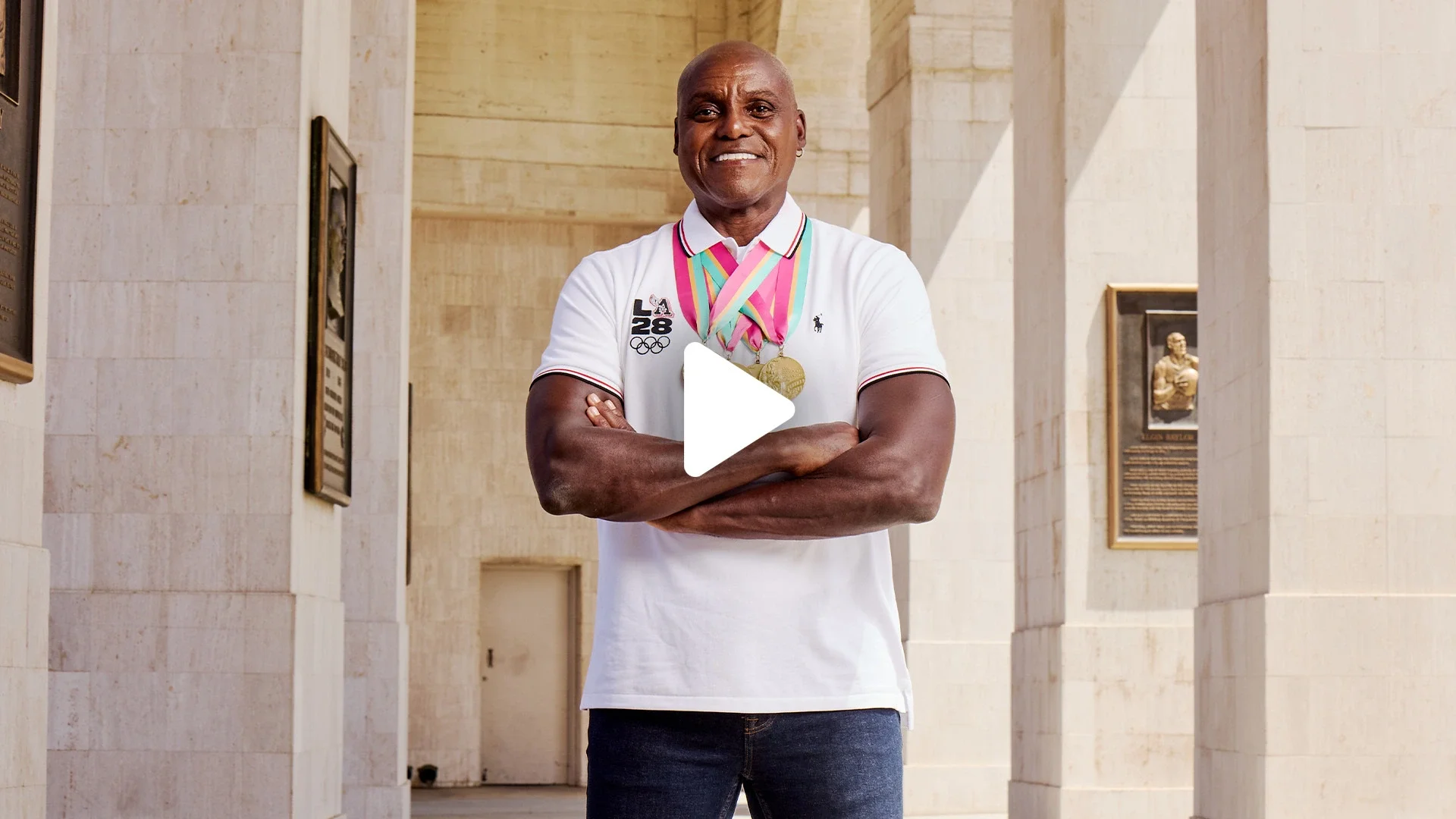 A photograph of Carl Lewis standing at the LA Memorial Coliseum wearing a white LA28 polo and wearing his four Olympic Gold medals around his neck