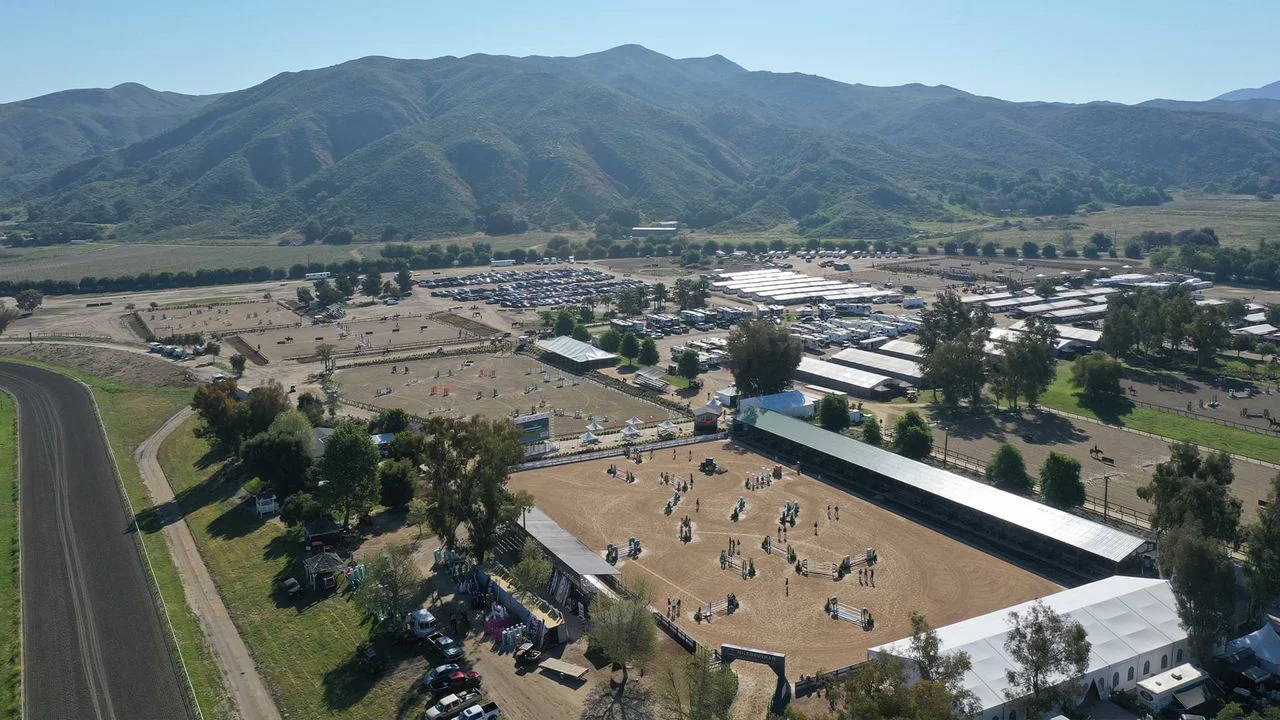 An aerial photograph of Galway Downs equestrian center showing five dirt horse arenas surrounded by trees, grass and various buildings as well as a few parking areas full of cars. The background shows rolling green hills full of grass, trees and bushes and a blue sky.