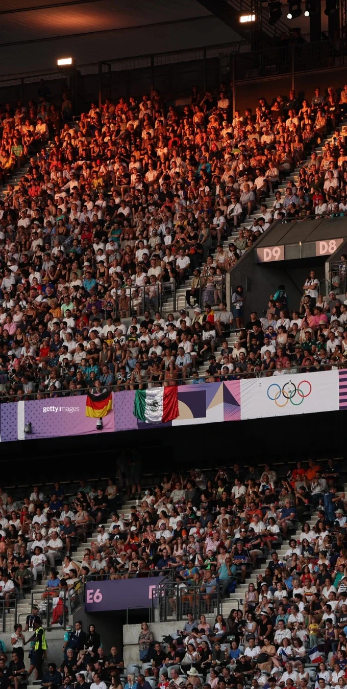 Fans in the stands at an indoor stadium at the Paris 2024 Olympic Games. The flags of Germany and Mexico hang from one of the railings next to signage of the Olympic rings.