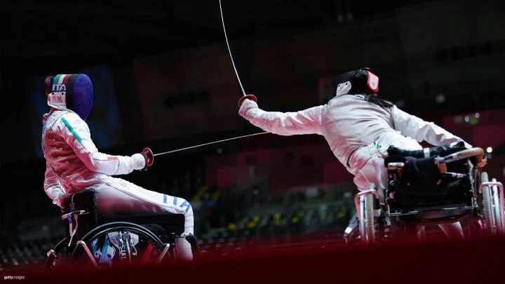 Two wheelchair fencers in action. Both athletes are dressed in full fencing attire, including masks and white protective clothing. The fencer on the left, with "ITA" and the Italian flag on their mask, is facing the fencer on the right, who is extending their arm and foil toward their opponent. The scene is set in an indoor arena with dim lighting and blurred background. 