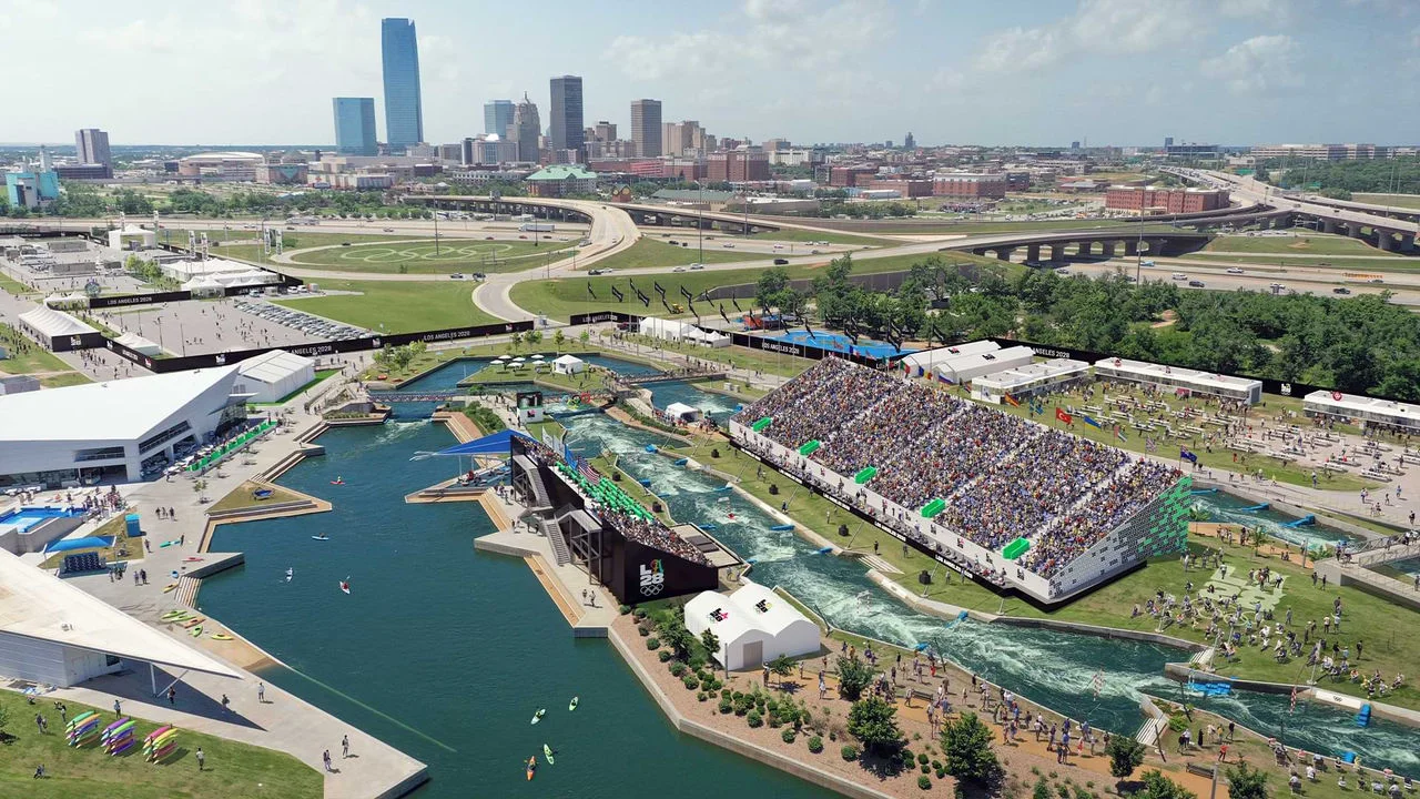 Una representación digital muestra el Whitewater Center también conocido como Riversport OKC. La imagen incluye un gran curso de agua con kayakistas navegando por los rápidos. Hay tribunas llenas de espectadores viendo el evento. Los alrededores cuentan con varios edificios, carpas y caminos con el logotipo de LA28. En el fondo, se ve el horizonte de la ciudad de Oklahoma City con edificios altos y carreteras. Los carteles del evento dicen "Los Angeles 2028", indicando que es una sede olímpica.