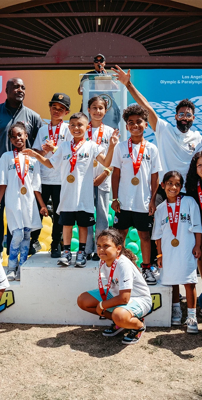 A group photo of multiple youth attendees from the LA28 Day of Sport, they are wearing white tshirts with the LA28 emblem on them