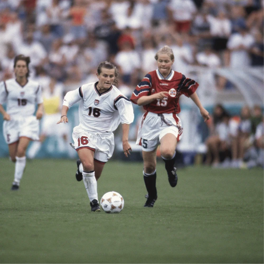 Tiffeny Milbrett corriendo tras un balón de Fútbol en un campo de césped, vistiendo una camiseta blanca de la selección femenina de Estados Unidos (USWNT) con el número 16, pantalones cortos blancos, medias blancas y tacos negros contra una jugadora de Noruega, con aficionados y otras jugadoras de fondo.