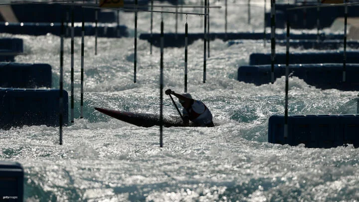 A canoer paddles through a turbulent white water course, navigating around hanging striped poles. The Canoe Slalom athlete, wearing a helmet, is silhouetted against the churning waves.