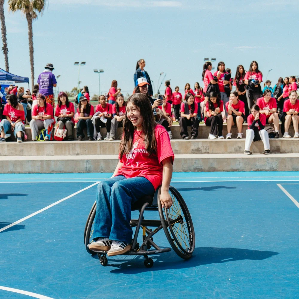 A photograph of a young girl laughing and smiling while sitting in a wheelchair on a tennis court