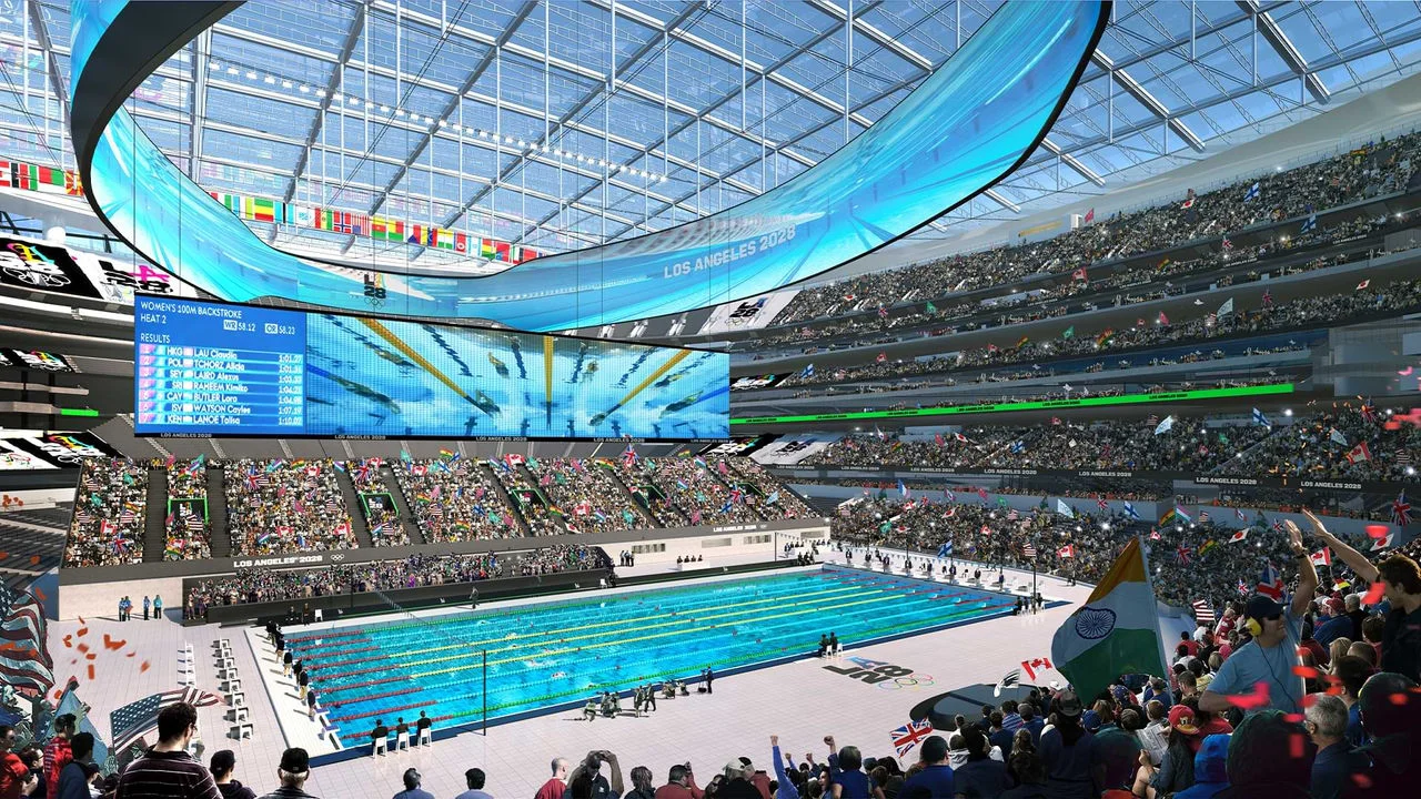 Representación del 2028 Olympic and Paralympic Stadium, también conocido como SoFi Stadium. El moderno recinto cubierto está lleno de aficionados con banderas de diferentes países. El enfoque está en la piscina, donde los atletas están compitiendo. Grandes pantallas sobre la piscina muestran el evento de los 100 metros espalda femenino e imágenes en vivo de las nadadoras. El estadio tiene un techo transparente y muchos niveles de asientos, todos llenos de espectadores.