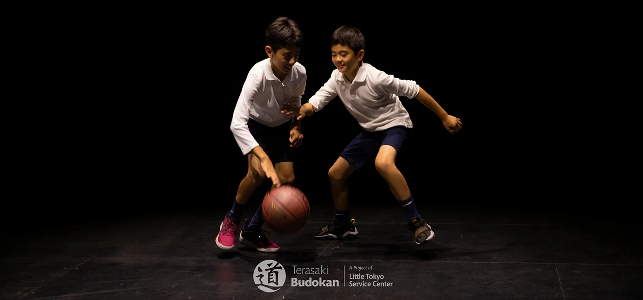 two young Japanese boys playing basketball one on one wearing long sleeved white polos and blue shorts, one boy is dribbling and the other is blocking and the text says Terasaki Budokan A Project of Little Tokyo Service Center