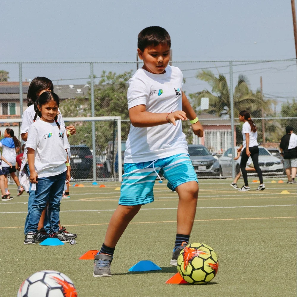A young boy kicking a soccer ball with a girl playing soccer behind him. Green grass with painted white lines and orange soccer cones is shown in the foreground.