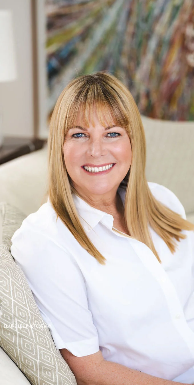 Photo of Maria Arena Bell, a caucasian female with shoulder length blond hair and bangs, sitting on a tan couch smiling and wearing a white collared shirt