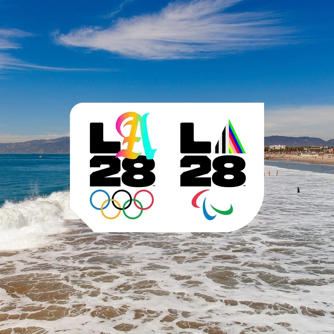 Ocean waves with sand in the forefront of the photograph with LA28 dual emblem superimposed on top.