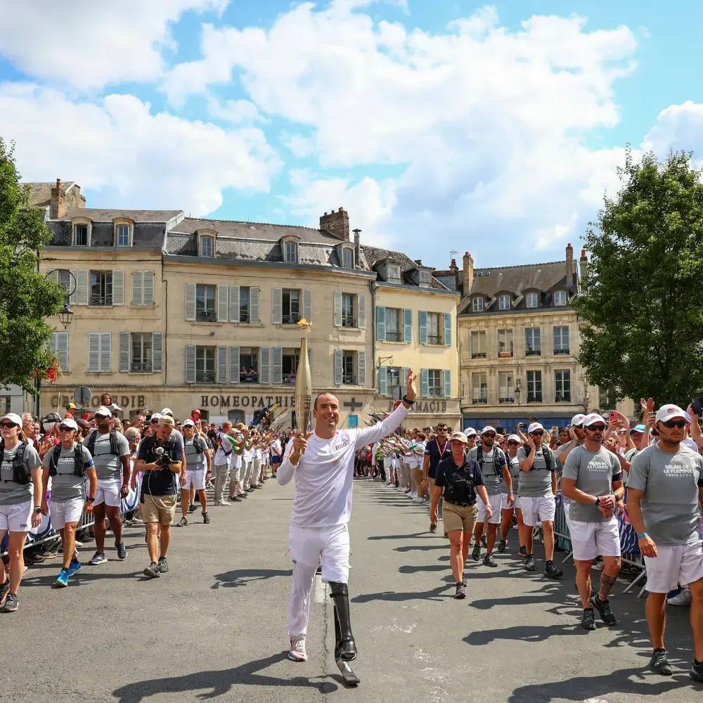 Man with a prosthetic leg running in a white long sleeve shirt and white shorts while carrying the Paralympic flame with a cityscape in the background