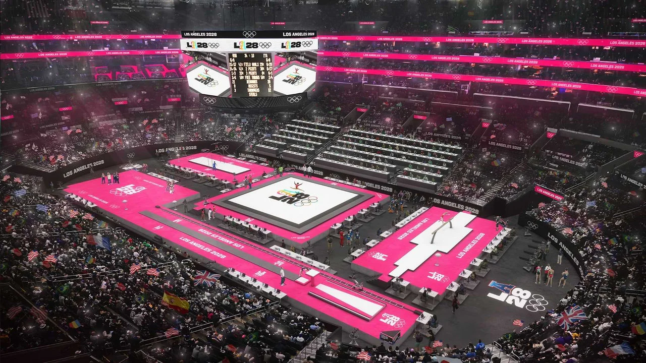 Representación del Los Angeles 2028 Olympic and Paralympic Games Arena, también conocido como el Crypto.com Arena. El estadio está lleno de espectadores, con áreas de asientos mostrando carteles vibrantes en color rosa con el texto "LOS ANGELES 2028" en texto blanco. En el piso están instalados varios aparatos de gimnasia, incluyendo la barra de equilibrio, las barras asimétricas y el área de ejercicio de suelo, todos con piso de color rosa. Atletas y oficiales son visibles cerca de los aparatos. Grandes pantallas digitales muestran el logo del evento, las puntuaciones y la información del evento. El público ondea varias banderas internacionales.