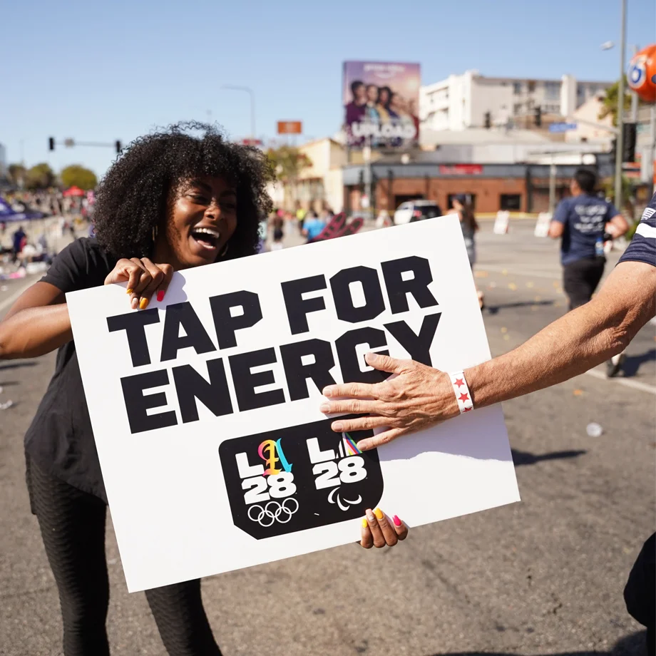 LA28 employee Corine Taylor smiling and holding up a sign that says Tap For Energy and someone is tapping the sign with their hand