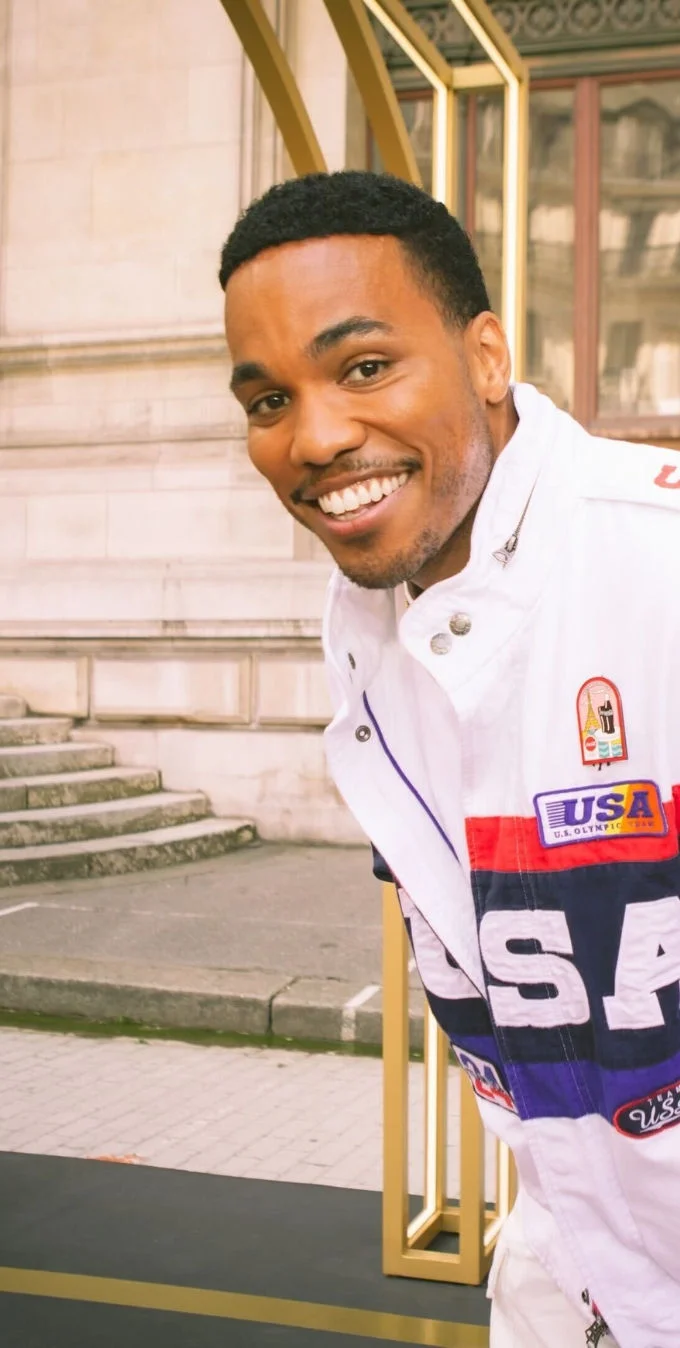 Una fotografía de Anderson .Paak sonriendo mirando a la cámara con un edificio de la ciudad detrás de él mientras lleva una chaqueta blanca Team USA Ralph Lauren Paris 2024 Ceremonia de Clausura.