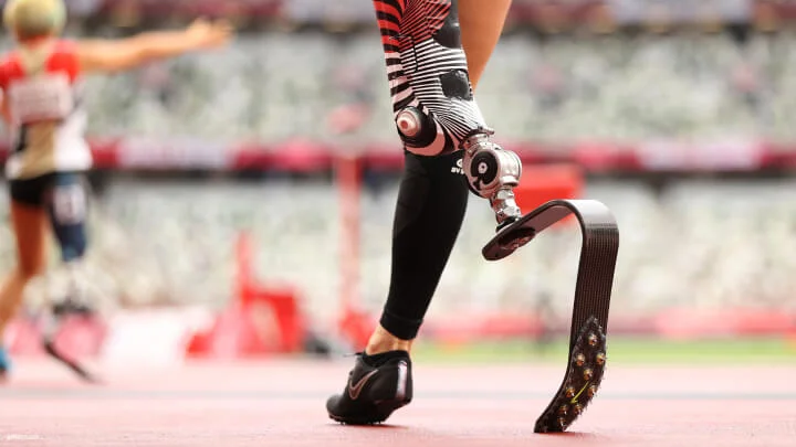 A close-up shot of a sprinter's left prosthetic running blade on a track. The prosthetic leg socket has a black, red, and white striped design, the blade is made of carbon fiber, and the athlete is wearing black running shoes and a black leg sleeve on their right leg. In the blurred background, another athlete with a prosthetic leg is visible.