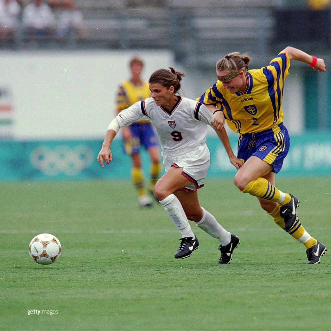 U.S. Women's Soccer National Team member Mia Hamm wearing white soccer socks, Nike cleats, a white jersey and white shorts with the number 9 on it while going after a ball on a grass field playing at the Atlanta 1996 Summer Games against Cecilia Sandell of Team Sweden wearing a printed bandana, yellow jersey and blue shorts with blue soccer socks and Nike cleats. 