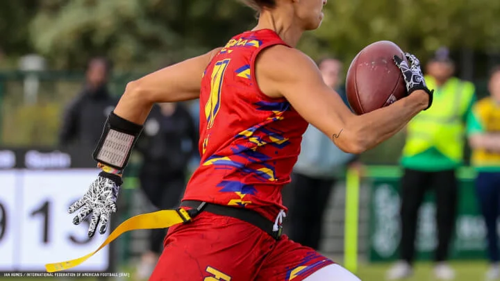 A close-up image shows a flag football player in action. The player is wearing a red sleeveless jersey with yellow and blue geometric patterns, white patterned gloves, and a black wristband with a playbook attached to their left wrist. They are holding a football in their right hand while running. A yellow flag is hanging from their waist, indicating a flag football game. The background shows blurred spectators and officials.