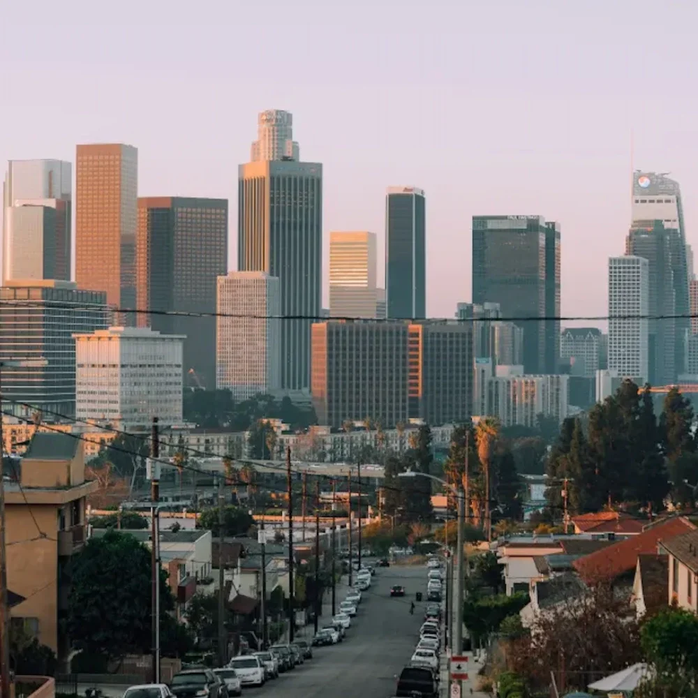 Downtown LA skyline as soon at sunset from a street on a hill lined with cars and apartment buildings. Trees and telephone poles can also be seen in the image.