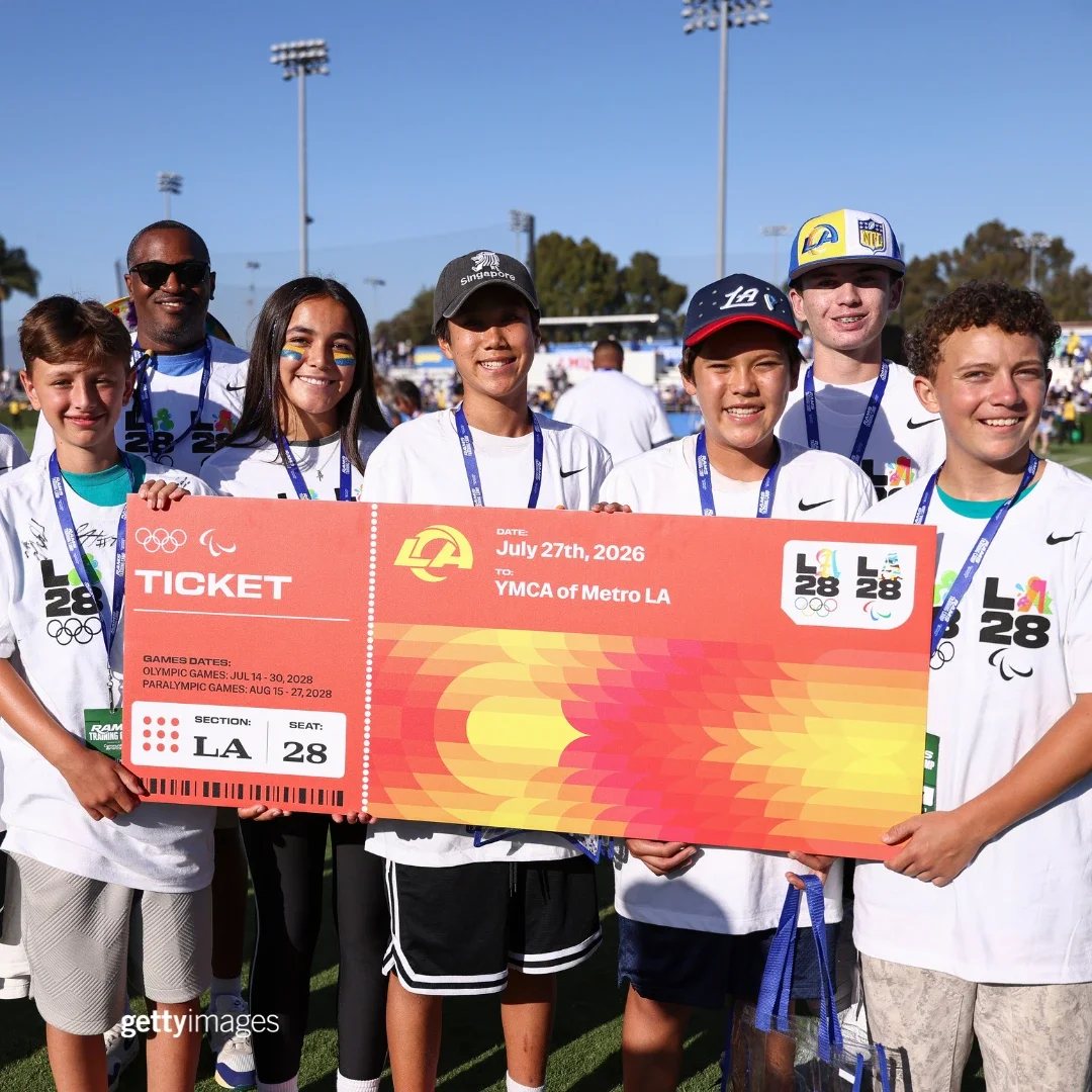 Children in white t-shirts holding up a LA28 big ticket sign in front of a football field 