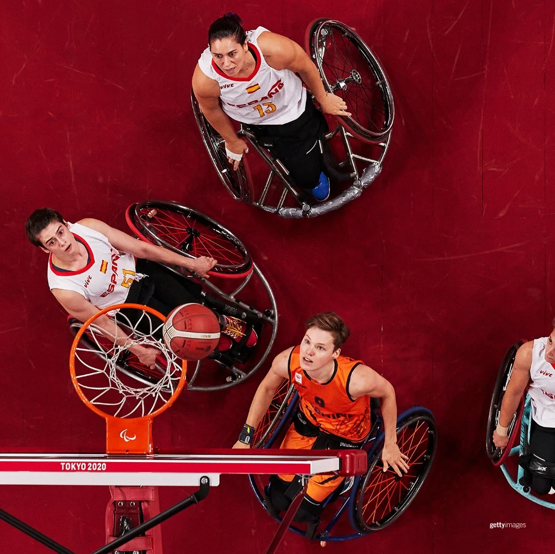 Paralympic wheelchair basketball players at the Tokyo 2020 Games in white Spain and orange Dutch basketball uniforms looking at the basketball hit the rim