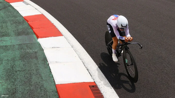 A cyclist wearing a Team USA uniform and an aerodynamic white helmet leans forward on a sleek black racing bike, navigating a curve on a race track. The track consists of dark gray asphalt and features sections with green asphalt and red-and-white curbs.