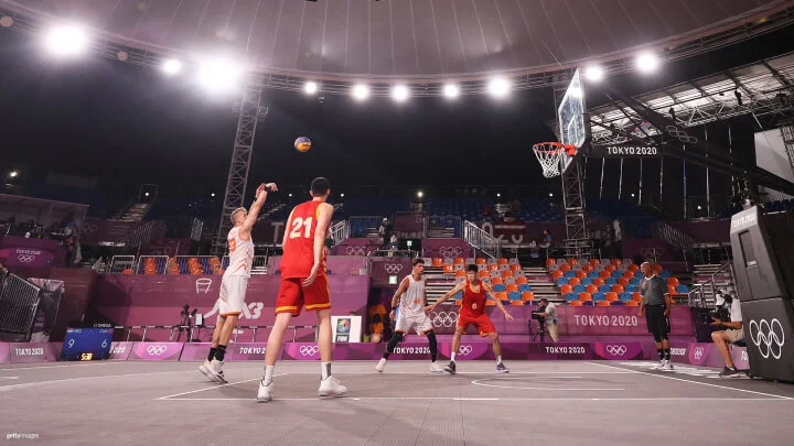 Un jugador de baloncesto con una camiseta gris está saltando intentando anotar punto, sosteniendo un balón azul y amarillo cerca del aro. Otro jugador al frente está defendiendo con un brazo levantado. Un tercer jugador está detrás, también intentando bloquear el tiro. Al fondo se ve una audiencia sentada en un estadio con algunas personas usando mascarillas.