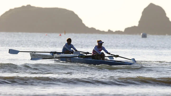 Dos remeros están en botes individuales en el océano, remando uno al lado del otro. El remero en primer plano lleva una camiseta blanca y roja, mientras que el remero en segundo plano viste una camiseta blanca y azul. Uno de los botes tiene el número de registro "GBR04" visible. Al fondo se ven islas rocosas, y el agua está ligeramente agitada.