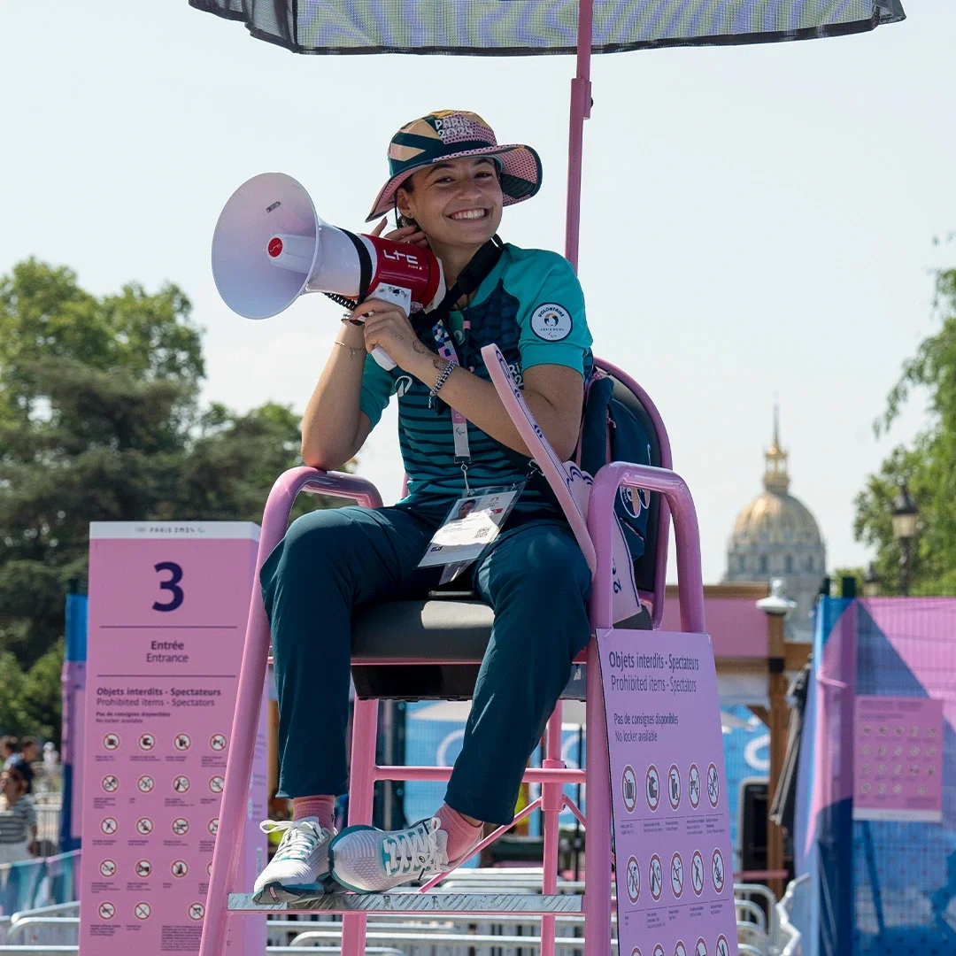 A Volunteer sitting in a chair with umbrella and microphone directing fans at the Paris 2024 Summer Games