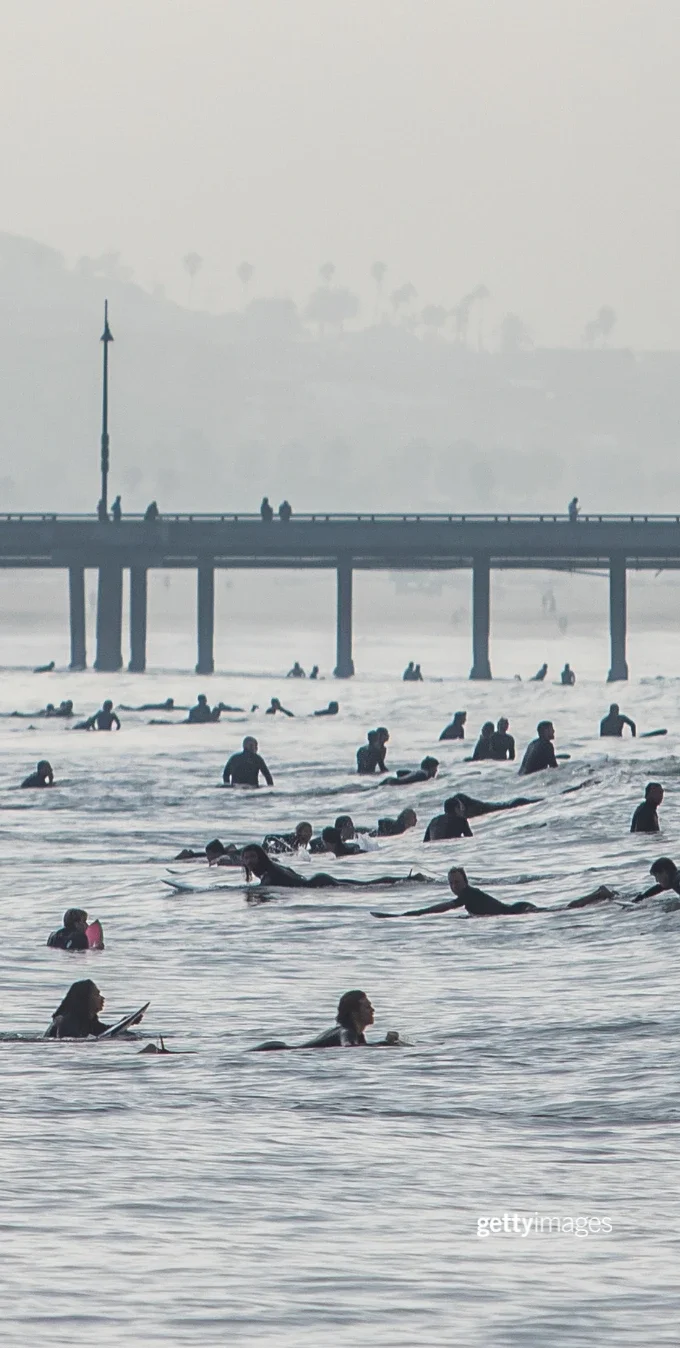 Surfers swim in the Pacific Ocean with a boardwalk in the background.