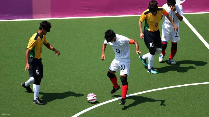 Four blindfolded soccer players are on a green artificial turf field, playing a match. Two players in yellow, green and black uniforms, wearing numbers 9 and 10, are interacting with two players in white uniforms with red socks, wearing numbers 10 and 11. The player in the white uniform with number 10 is dribbling a white and pink soccer ball. The background features a pink and purple banner along the edge of the field.