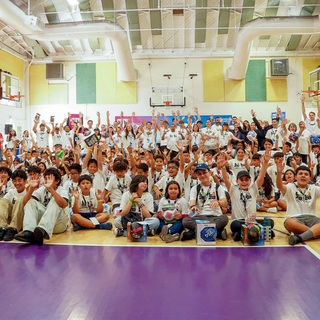 Children raising their hands inside a gymnasium while wearing white LA28 t-shirts during the 2026 LA28 Day of Sport. 
