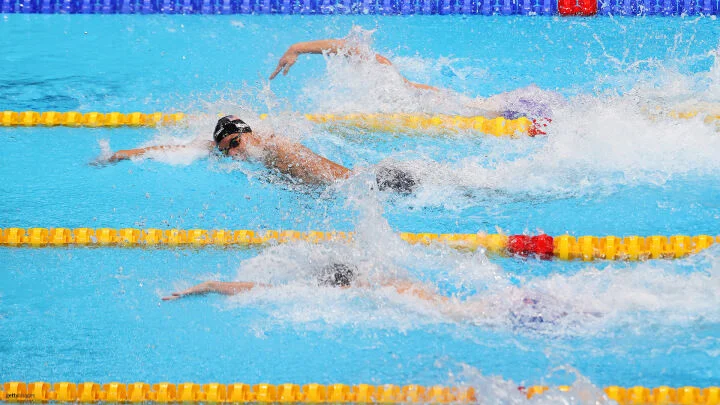 Three swimmers are competing in a freestyle race, swimming side by side in separate lanes. The swimmer in the center lane is slightly ahead of the others. The water is clear and blue, with splashes around the swimmers as they move. Yellow lane dividers separate the lanes.