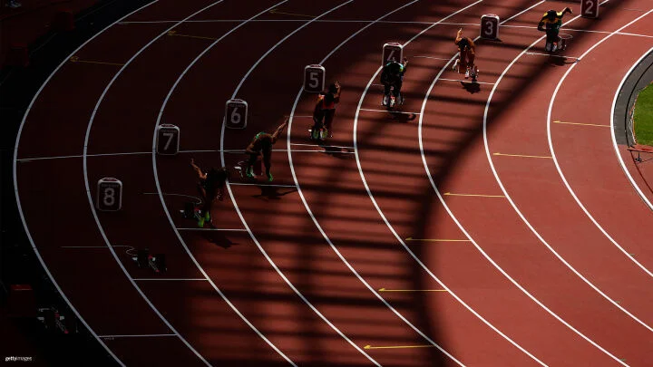 A group of sprinters are captured at the starting blocks of a race on a curved track under dramatic lighting. The shadows from the stadium structure create a pattern across the track. Each runner is in the starting block, launching forward. Lane numbers from 2 to 8 are visible.