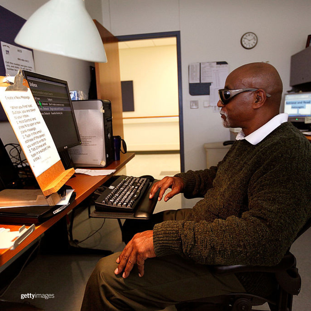 A man wearing glasses looks at a computer screen with a screenreader to his left. 