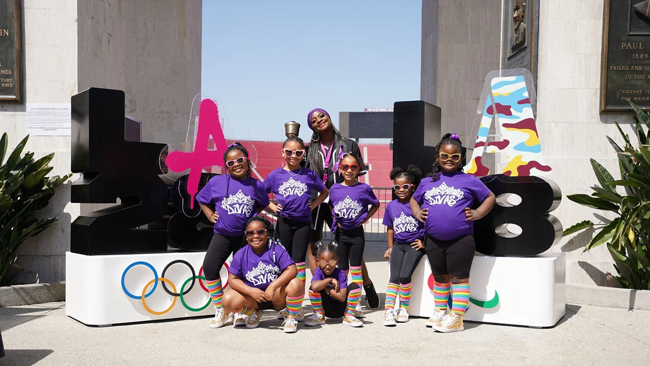 Group photo of seven young African American girls smiling wearing purple shirts that says Divas, black leggings and rainbow socks, standing in front of LA28 logos