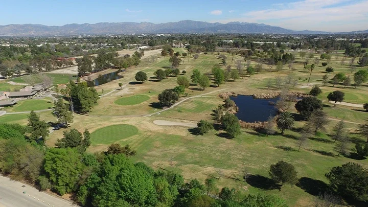 Representación del Sepulveda Basin Recreation Area preparado para los Juegos Olímpicos y Paralímpicos de Los Ángeles 2028. El área cuenta con múltiples recintos deportivos con tribunas llenas de espectadores. Los recintos incluyen una pista de BMX con una rampa elevada y un parque de patinaje con varias rampas y obstáculos. Alrededor de los recintos deportivos hay espacios verdes. En primer plano se ve un gran lago con una pequeña cascada. El fondo muestra un paisaje urbano con montañas. Banderas de "Los Angeles 2028" y decoraciones temáticas olímpicas son visibles en toda el área.